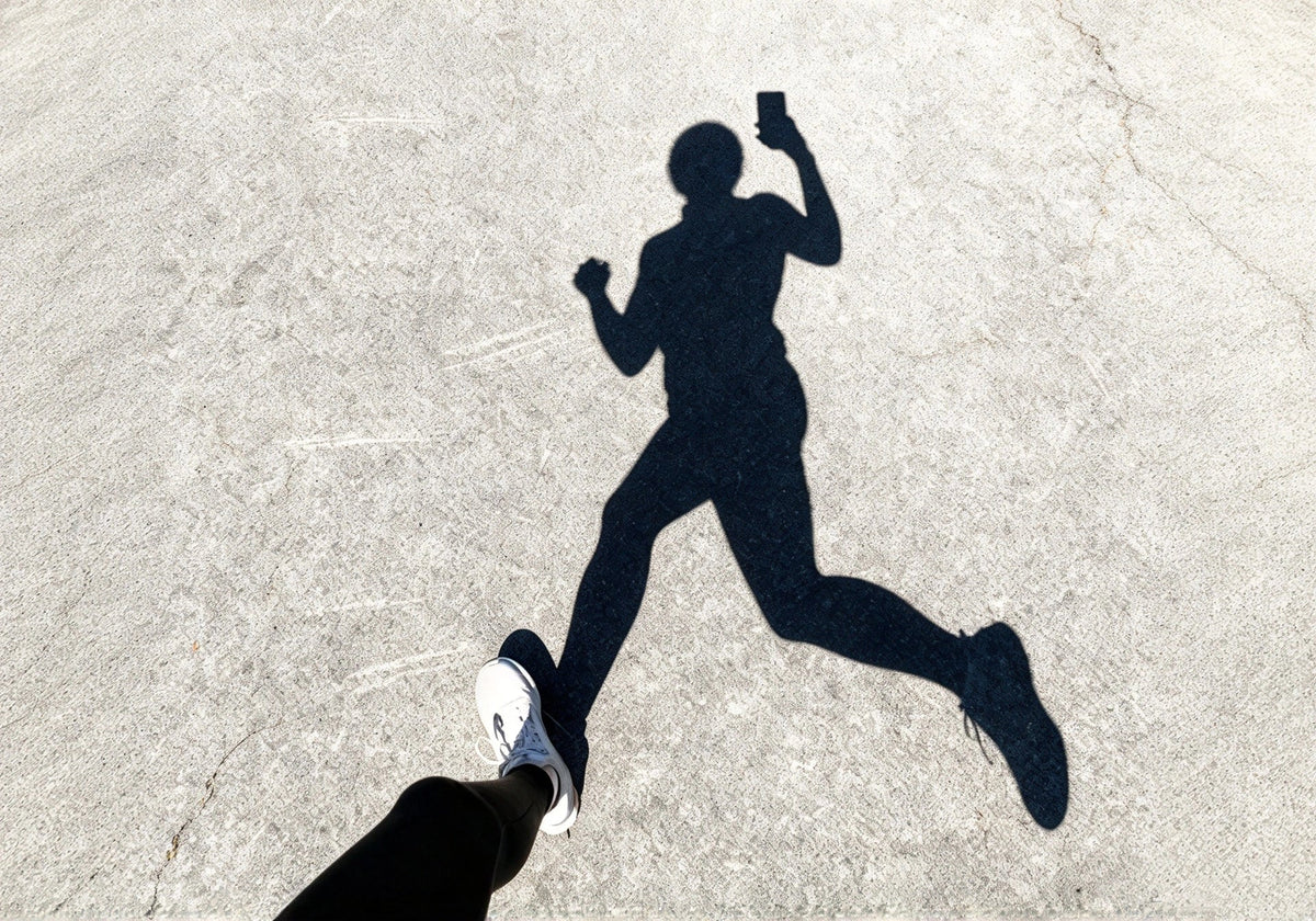 A runner's shadow cast on the pavement while holding a phone during a daytime workout.